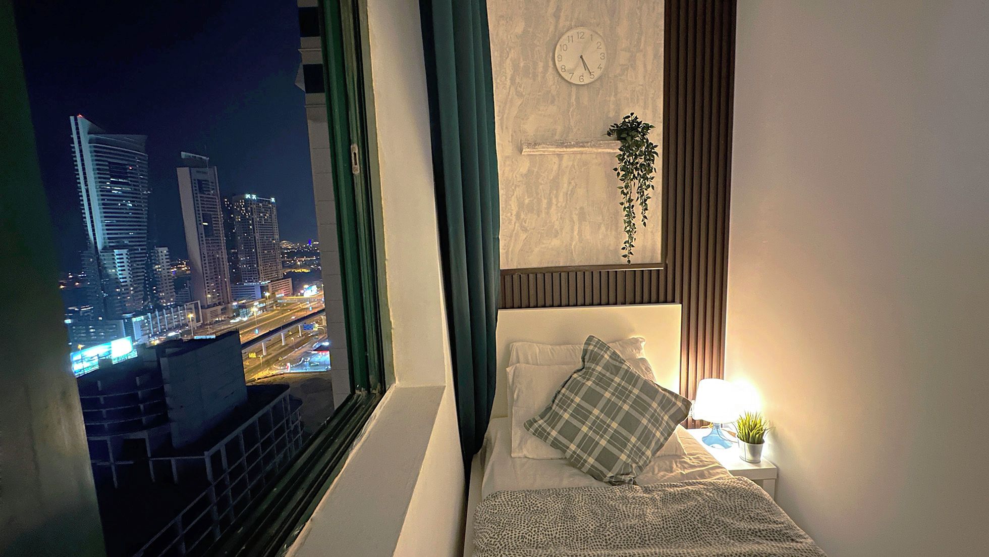 Een slaapkamer met uitzicht op de stad, een klok, een kamerplantje en een lamp.