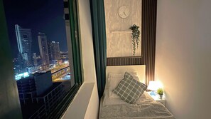 A bedroom with a city view, a clock, a bedside plant, and a lamp.