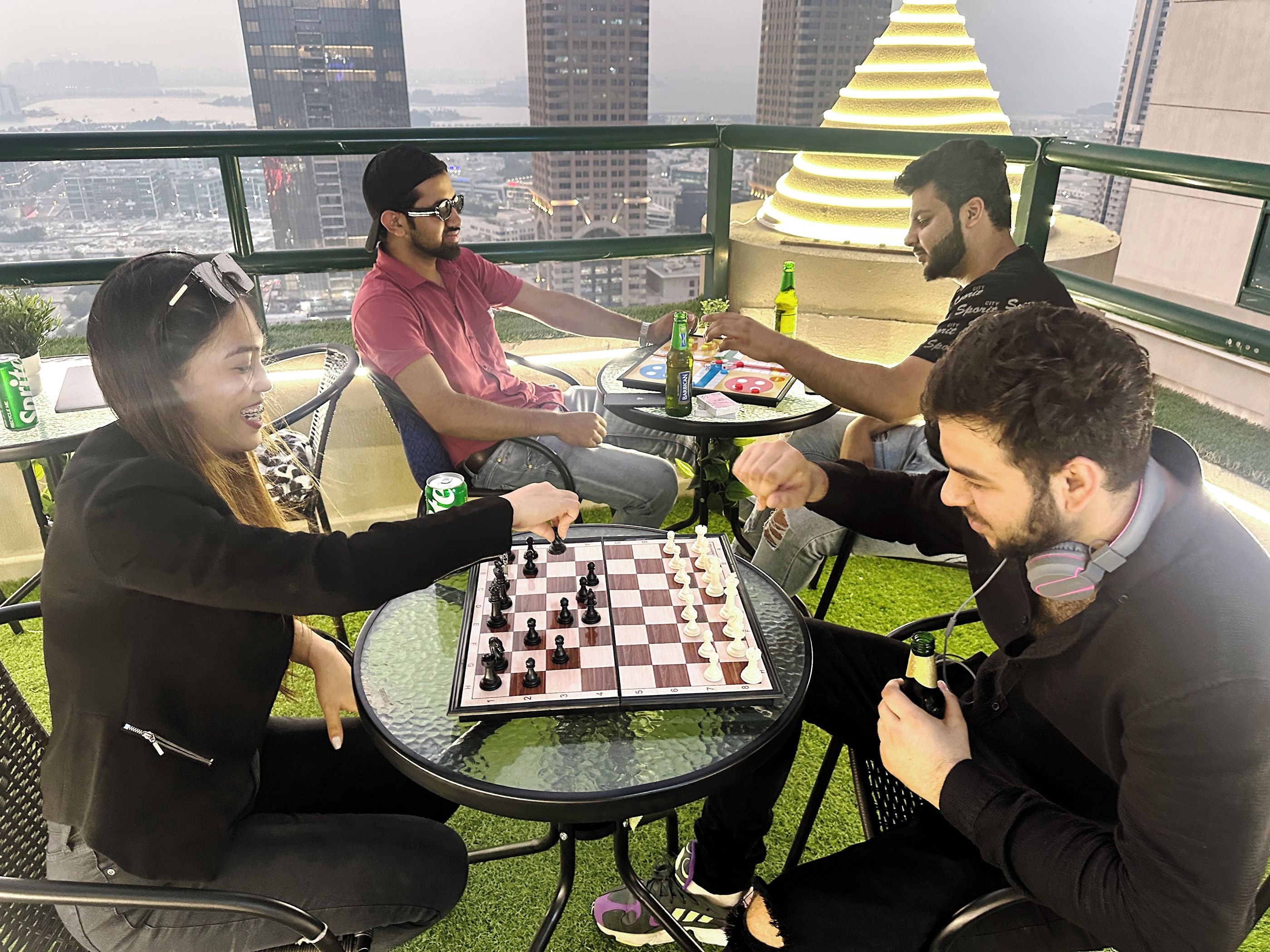 A group of people playing chess on a glass-top table with a city view in the background.
