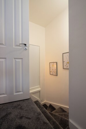 A hallway with a white door, a staircase with dark carpeting, and two framed pictures on the wall. - Bright Mezzanine One Bedroom Central Apartment (Edinburgh)