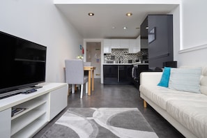 Una sala de estar moderna con un televisor de pantalla plana, un sofá con cojines, un área de comedor con una silla y una cocina con armarios y salpicadero.