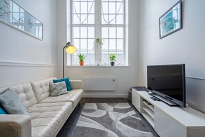 A modern living room with a sofa, a TV, a lamp, and a window with plants. - Bright Mezzanine One Bedroom Central Apartment (Edinburgh)