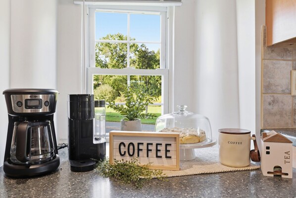 A kitchen with a coffee maker, a mug, a sign reading 'COFFEE', and a view of greenery outside.