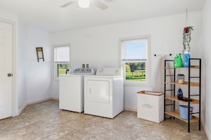 A laundry room with two washing machines, a dryer, a shelf with cleaning supplies, and a window with a view of a green landscape.