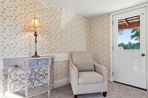 A room with floral wallpaper, a lamp on a side table, a chair with a cushion, and a white door with a window.