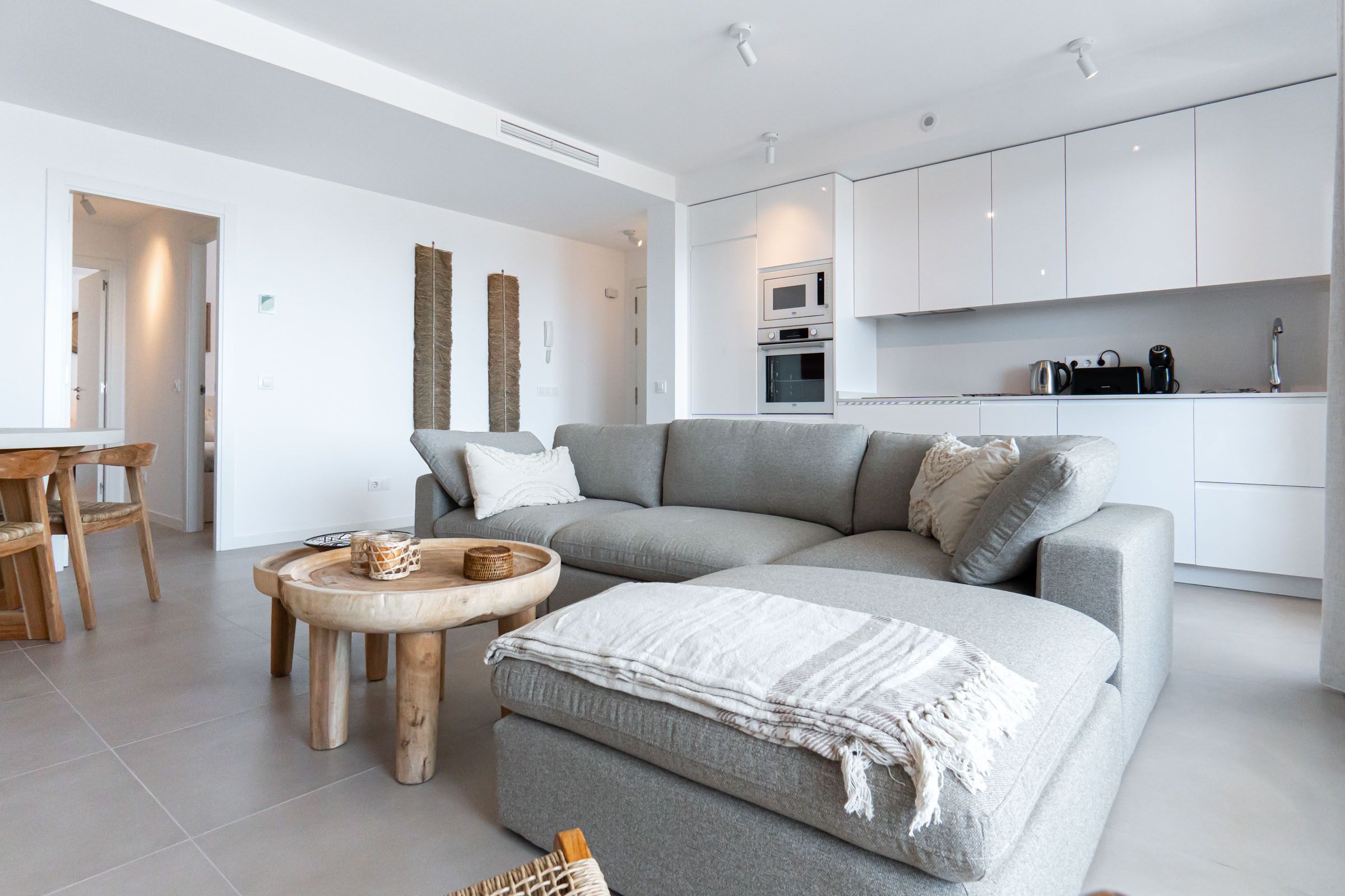 Un saló modern amb un sofà seccional gris, una taula de centre de fusta i una zona de cuina amb armaris i electrodomèstics blancs.