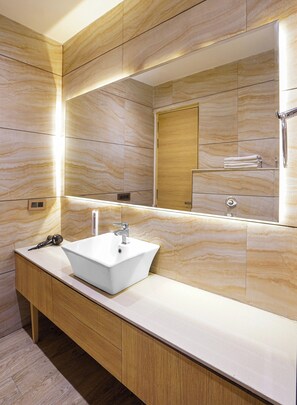 Un baño con un lavamanos blanco, tocador de madera, paredes de mármol y un espejo grande.