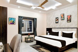 Habitación de hotel con cama, escritorio, silla, ventana con cortinas, ventilador de techo y decoración en la pared.