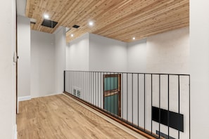 A hallway with wooden flooring, white walls, a black metal railing, and a door with a green frame. - The Chestnut House (Tomball)