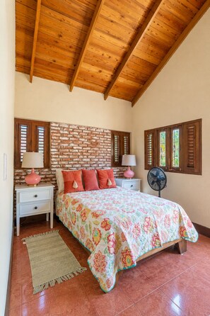 A bedroom with a brick wall, wooden ceiling beams, a bed with floral bedding, two bedside tables with lamps, and a fan. - Villa 13 Jarabacoa with AC/ Wifi Jacuzzi (Jarabacoa)