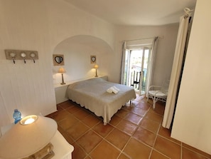 A bedroom with a bed, a bedside table, a lamp, a chair, and a view of the outdoors. - Li Conchi Villa 17 - detached villa within the Li Conchi Village (Castiadas)