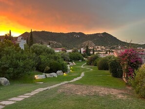 A garden pathway leading to a stone building with a traditional roof, surrounded by lush greenery and a mountainous backdrop. - Li Conchi Villa 17 - detached villa within the Li Conchi Village (Castiadas)