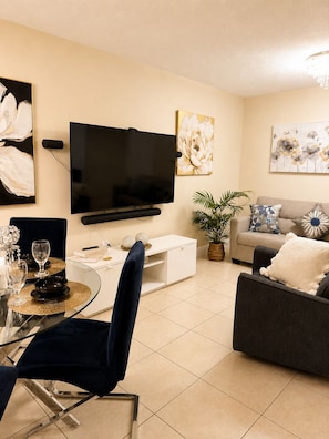 Ein modernes Wohnzimmer mit Flachbildfernseher, Essbereich mit Glastisch und einer Couch mit Zierkissen.