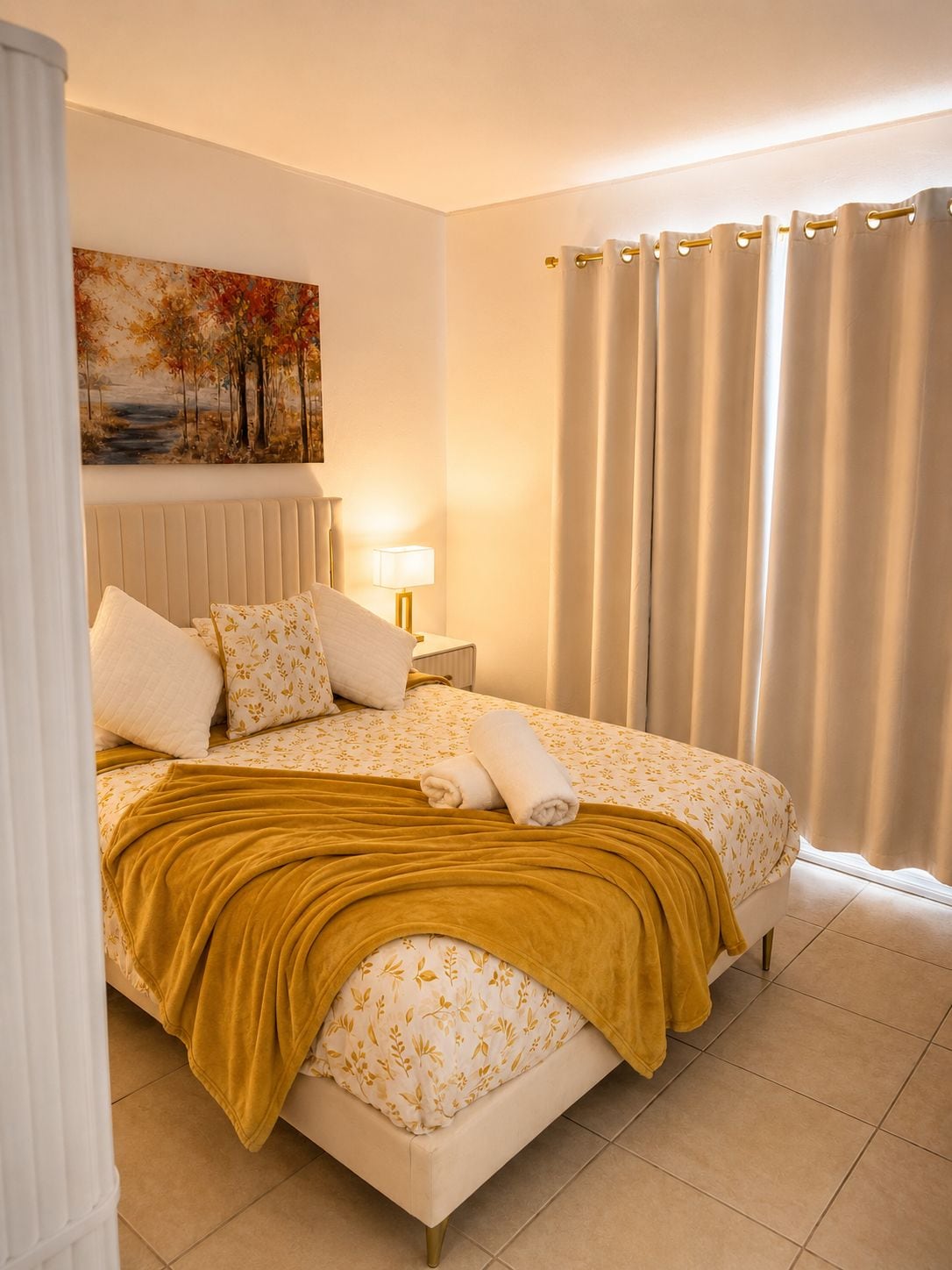Un lit bien fait, avec une couette à motifs floraux, un plaid de couleur moutarde et deux serviettes enroulées.