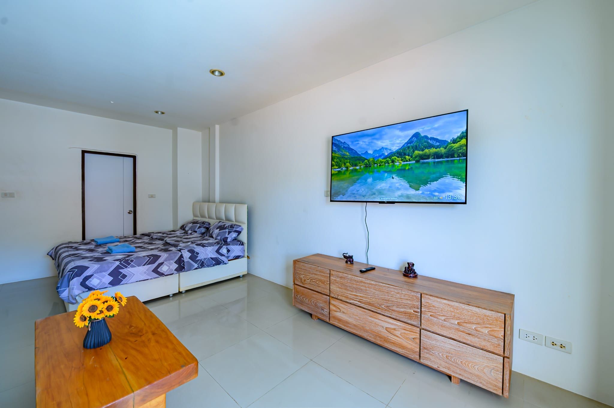 Un dormitorio con una cama, un aparador de madera, un televisor en la pared y un florero con flores sobre una mesa.