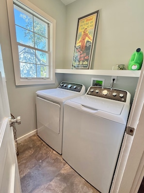 A laundry room with two front-loading washing machines, a window with a view of trees, and a poster on the wall. - Charming updated home w/large bedrooms & big deck. Great for groups & families! (Shelbyville)