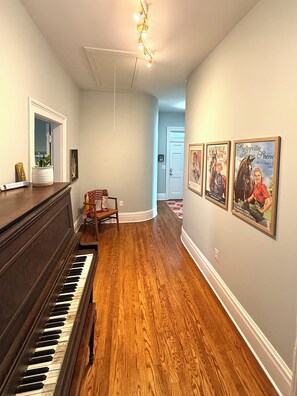 Un couloir avec un piano, des tableaux encadrés accrochés au mur et une porte au fond.