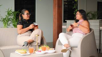 Two individuals seated on wicker chairs at an outdoor dining area with a table set with plates, glasses, and a teapot.
