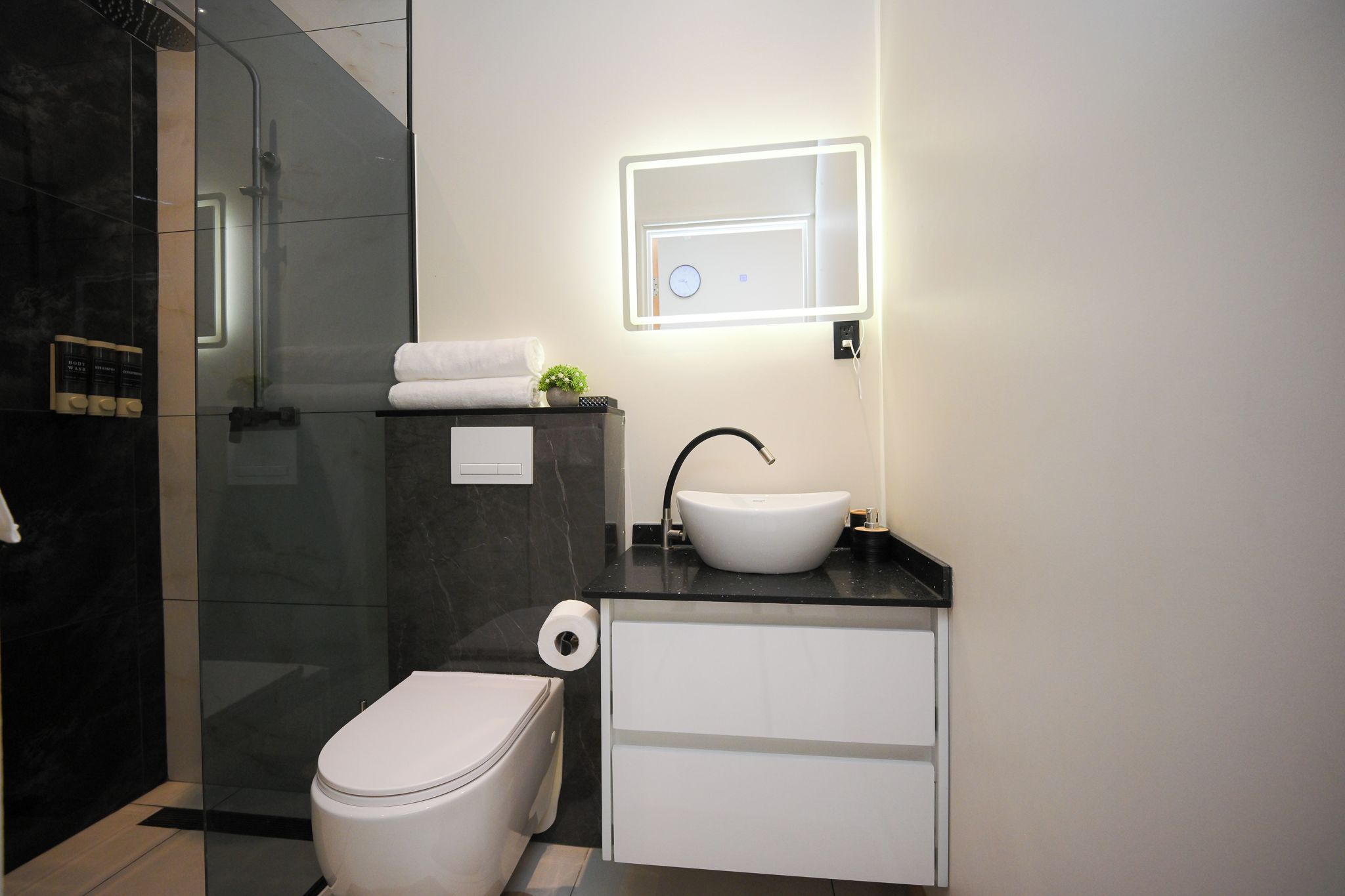 Une salle de bain moderne avec une douche à parois de verre, des toilettes blanches, un lavabo avec un robinet noir et un luminaire fixé au mur.