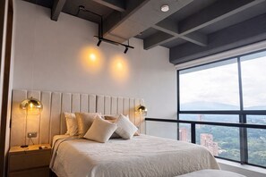 A modern bedroom with a large bed, two bedside lamps, a nightstand, and a city view through a large window. - Luxury Duplex with City Views (Medellín)