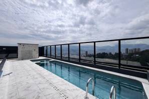 Una piscina al terrat amb vista a la ciutat, una petita construcció de pedra i una barana metàl·lica.