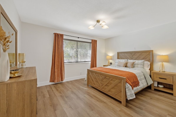 Un dormitorio con una cama de madera, una mesita de noche, una lámpara de noche, una ventana con cortinas y un espejo en la pared.