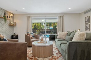 A living room with a sofa, armchair, and coffee table, a brick wall, and a view of a backyard with a pool.
