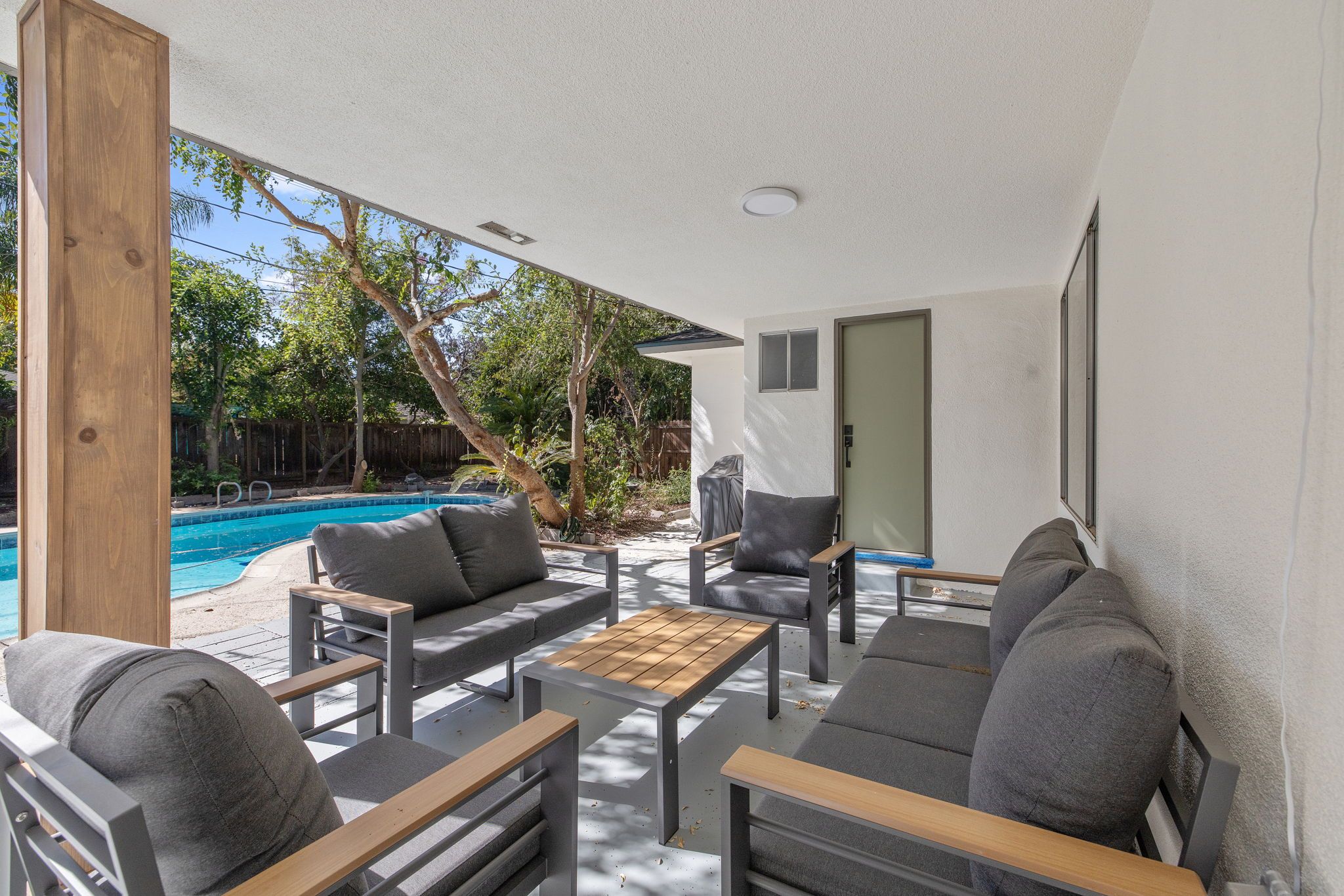 An outdoor patio with a pool, grey sofas, and wooden tables.