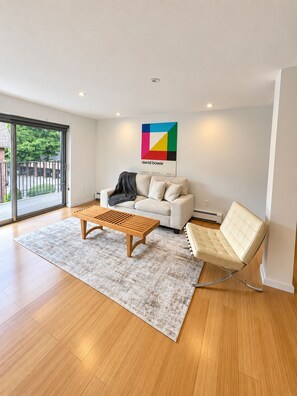 Ein modernes Wohnzimmer mit einer Couch, einem Holztisch und einem Ledersessel. An der Wand hängt ein farbenfroher Plakat und ein großes Fenster bietet einen Ausblick nach draußen.