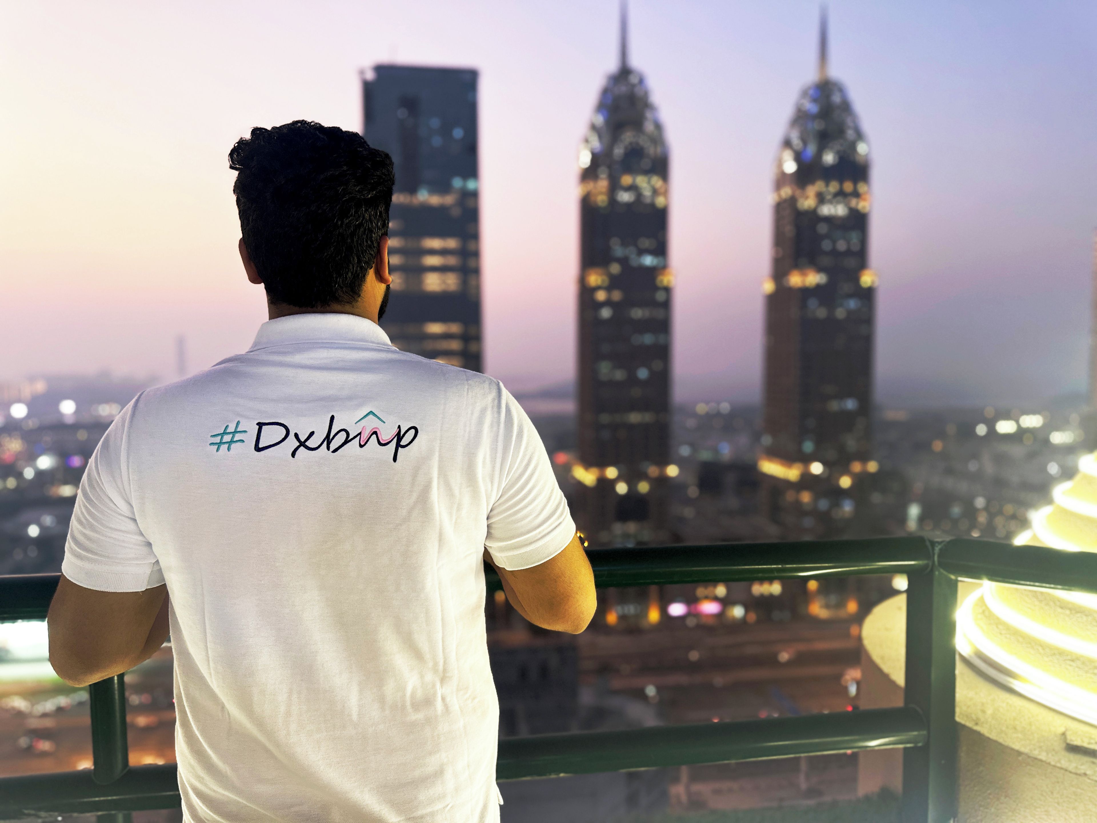 Een persoon in een wit shirt met een hashtag en een logo, kijkt uit over een stadsgezicht met twee opvallende wolkenkrabbers.