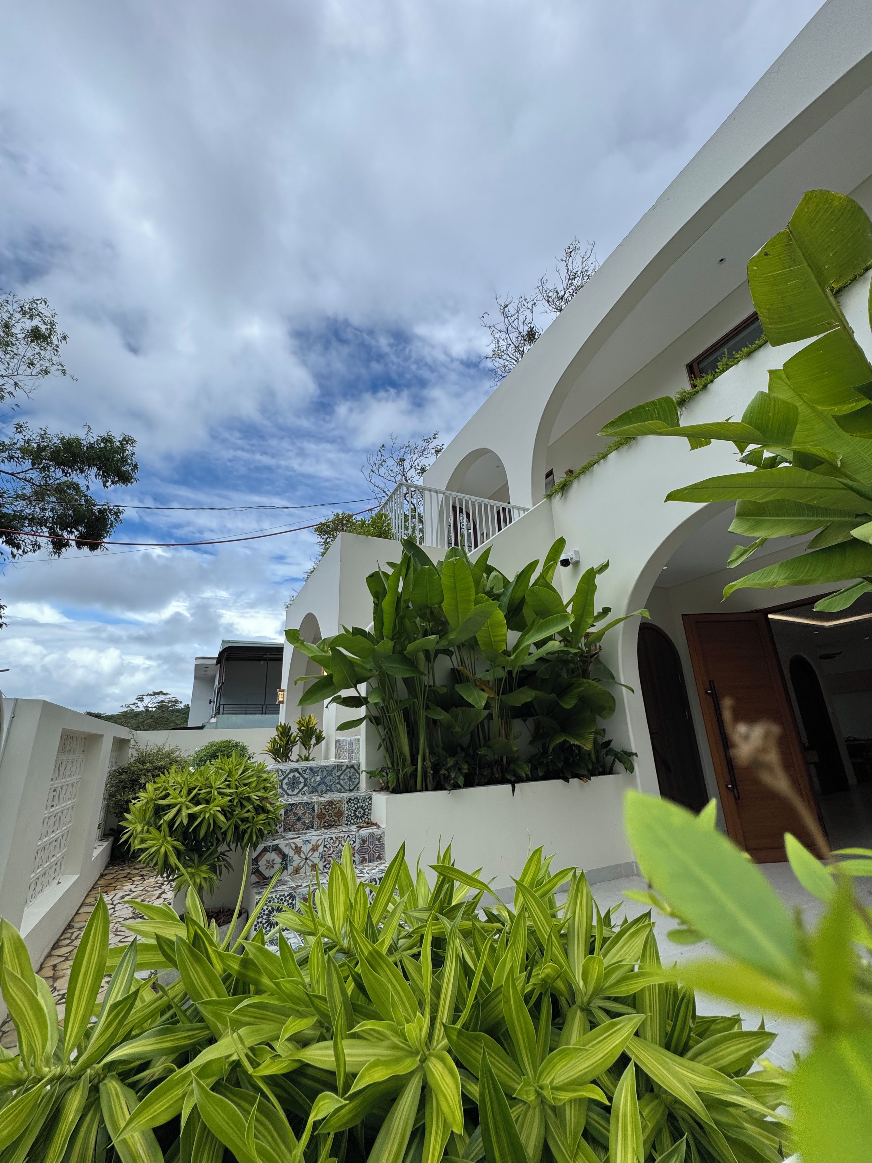 A white building with a balcony, surrounded by lush green plants.