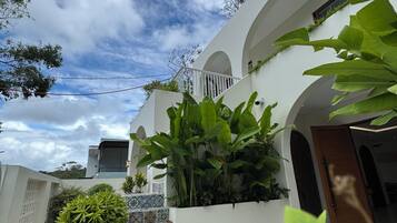A white building with a balcony, surrounded by lush green plants.