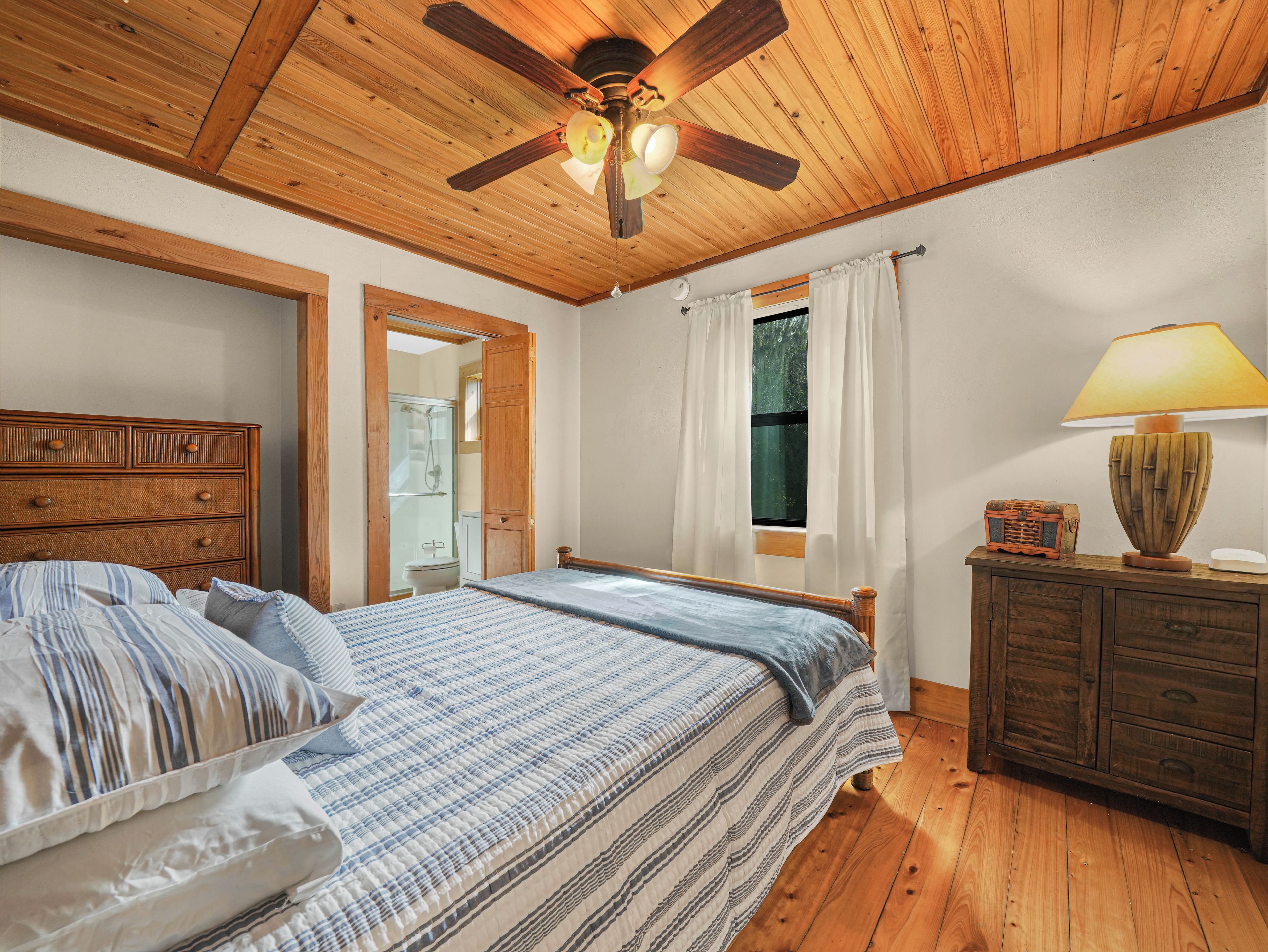 A bedroom with a wooden ceiling, a bed with striped bedding, a nightstand with a lamp, and a window with curtains.
