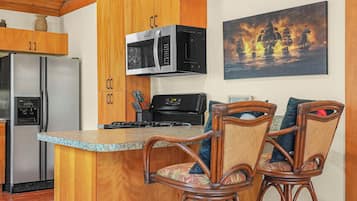 Una cocina con armarios de madera, un refrigerador de acero inoxidable, un microondas y una estufa. Hay dos sillas de mimbre con cojines.