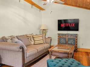A living room with a sofa, a glass-top coffee table, a television displaying 'NETFLIX', and a ceiling fan.