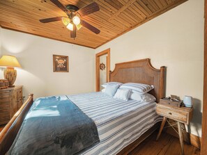 A bedroom with a wooden ceiling fan, a bed with striped bedding, a nightstand with a lamp, and a door with a ship's wheel decoration.