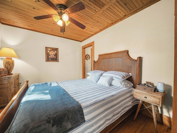 A bedroom with a wooden ceiling fan, a bed with striped bedding, a nightstand with a lamp, and a door with a ship's wheel decoration.