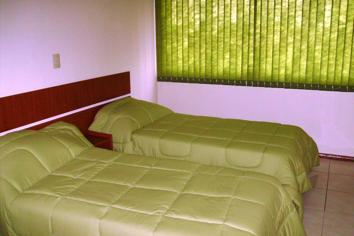 A room with two beds, a wooden headboard, a window with blinds, and a tiled floor.