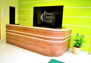 A hotel reception desk with a modern design, a flat-screen TV, and a potted plant. - Hotel Ouro Verde (Charqueadas)