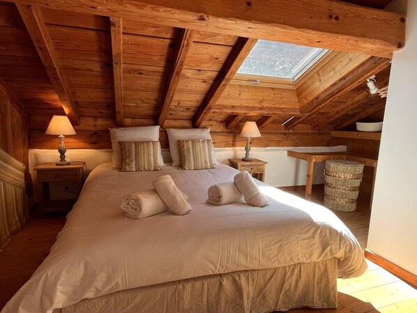 Une chambre avec un grand lit, des poutres apparentes en bois au plafond, une lucarne et des lampes de chevet.