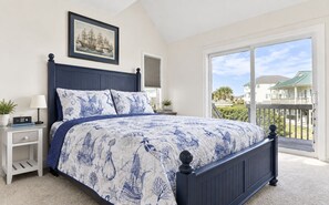 A bedroom with a large bed, a nightstand, a lamp, a framed picture on the wall, and a view of a balcony with a house in the background. - Nigh Noon - Ocean view, 50 yards to the beach (Surf City)