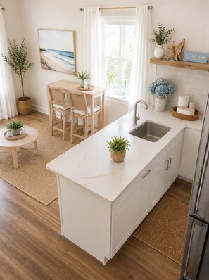 A modern kitchen with a central island, a dining area with a wooden table and chairs, and a view of the outdoors through a window. - Nigh Noon - Ocean view, 50 yards to the beach (Surf City)