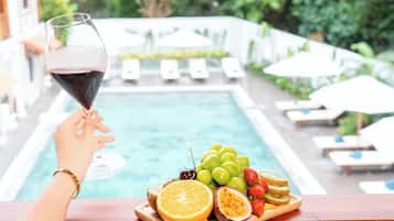 Vista de la piscina con una copa de vino tinto, una bandeja de madera con frutas y sillones de descanso.