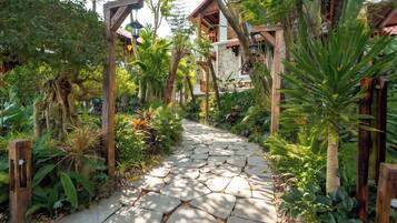 A stone pathway leading to a wooden structure with a thatched roof, surrounded by lush tropical vegetation.