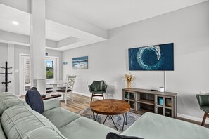 A modern living room with a sectional sofa, a dining area with a table and chairs, a large ocean-themed artwork on the wall, and a view of the outdoors through a door. - Steps to the Beach | Sleeps 13 | 2 Living Spaces (Port St Joe)