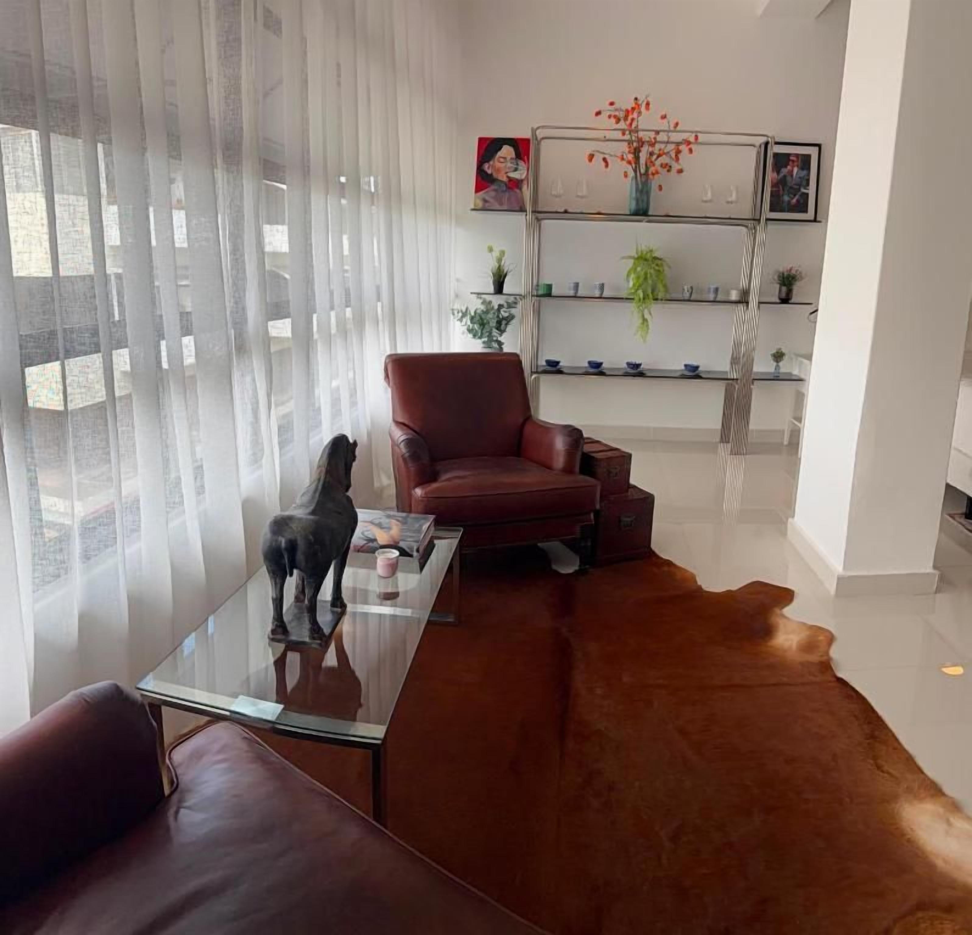 Una sala de estar moderna con una mesa de centro de vidrio, un sofá de cuero y un sillón rojo. Se observa una estantería con objetos decorativos y una planta.