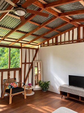 Ein Zimmer mit schrägem Dach, Holzbalken, einem Fernseher, einer Couch, einem Sessel, einem kleinen Tisch und einer Zimmerpflanze.
