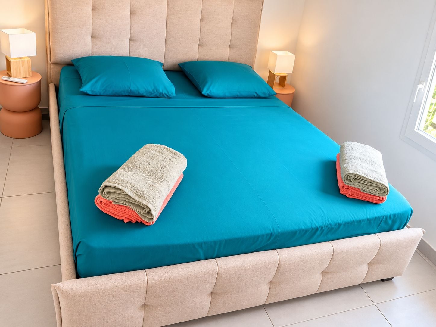 A bed with a teal bedspread, two pillows, and two rolled towels.