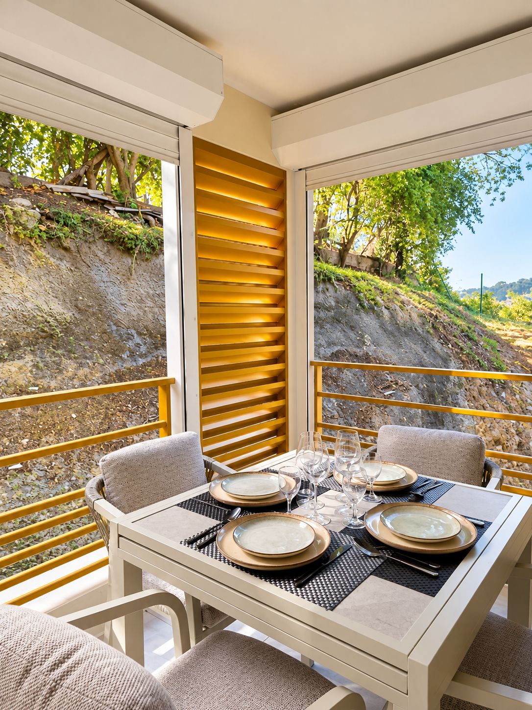 An outdoor dining area with a table set for four, surrounded by chairs, and overlooking a landscaped garden with a view of a hillside.