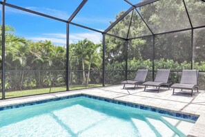 Ein Poolbereich mit freiem Blick auf die umliegende Bepflanzung, eine überdachte Sitzgelegenheit mit Stühlen und ein Pool mit Fliesenbordstein.